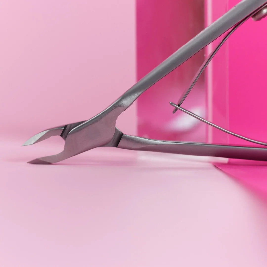 A close-up of a metallic nail clipper or cuticle tool resting diagonally on a pink surface with a matching pink background.