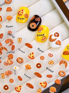 A close-up of decorative nail wraps or stickers displayed on a white padded tray, featuring illustrated food motifs such as pizza, bread, toast, croissants, cupcakes, and labels like 'Pizza,' 'Bread,' and 'Jelly.'.