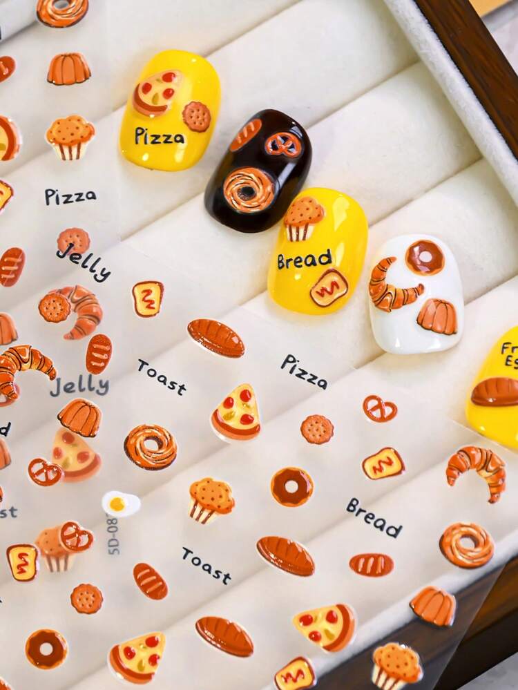 A close-up of decorative nail wraps or stickers displayed on a white padded tray, featuring illustrated food motifs such as pizza, bread, toast, croissants, cupcakes, and labels like 'Pizza,' 'Bread,' and 'Jelly.'.