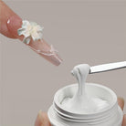 A close-up of a hand holding a white cosmetic jar while another hand displays a long nail decorated with a white flower and clear adhesive details. A metal spatula lifts thick white cream from the jar against a plain taupe background.