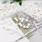 White square sugar cubes scattered on a white surface with two clear plastic containers holding more cubes, a silver metal tool, and printed paper props in the background.
