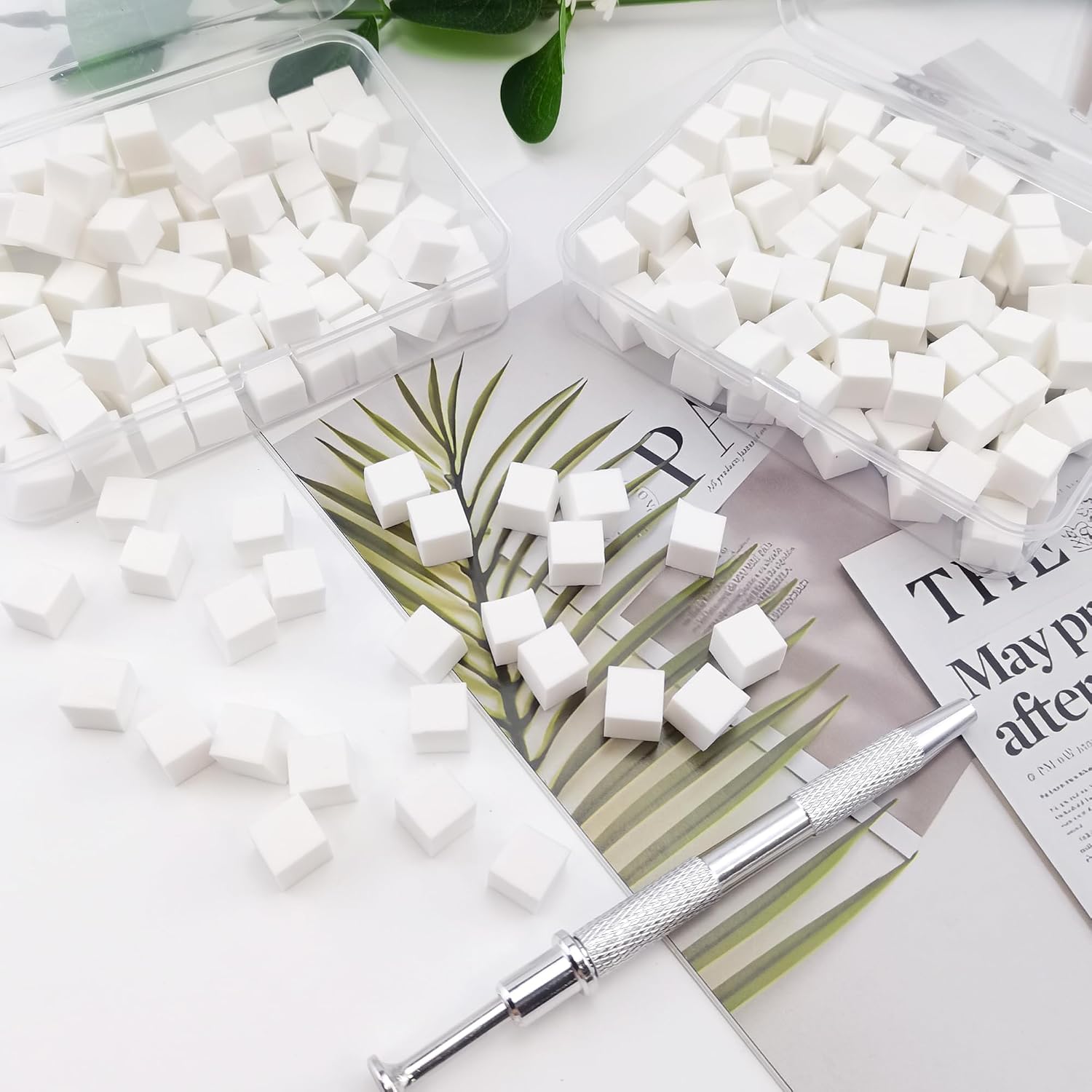 White square sugar cubes scattered on a white surface with two clear plastic containers holding more cubes, a silver metal tool, and printed paper props in the background.
