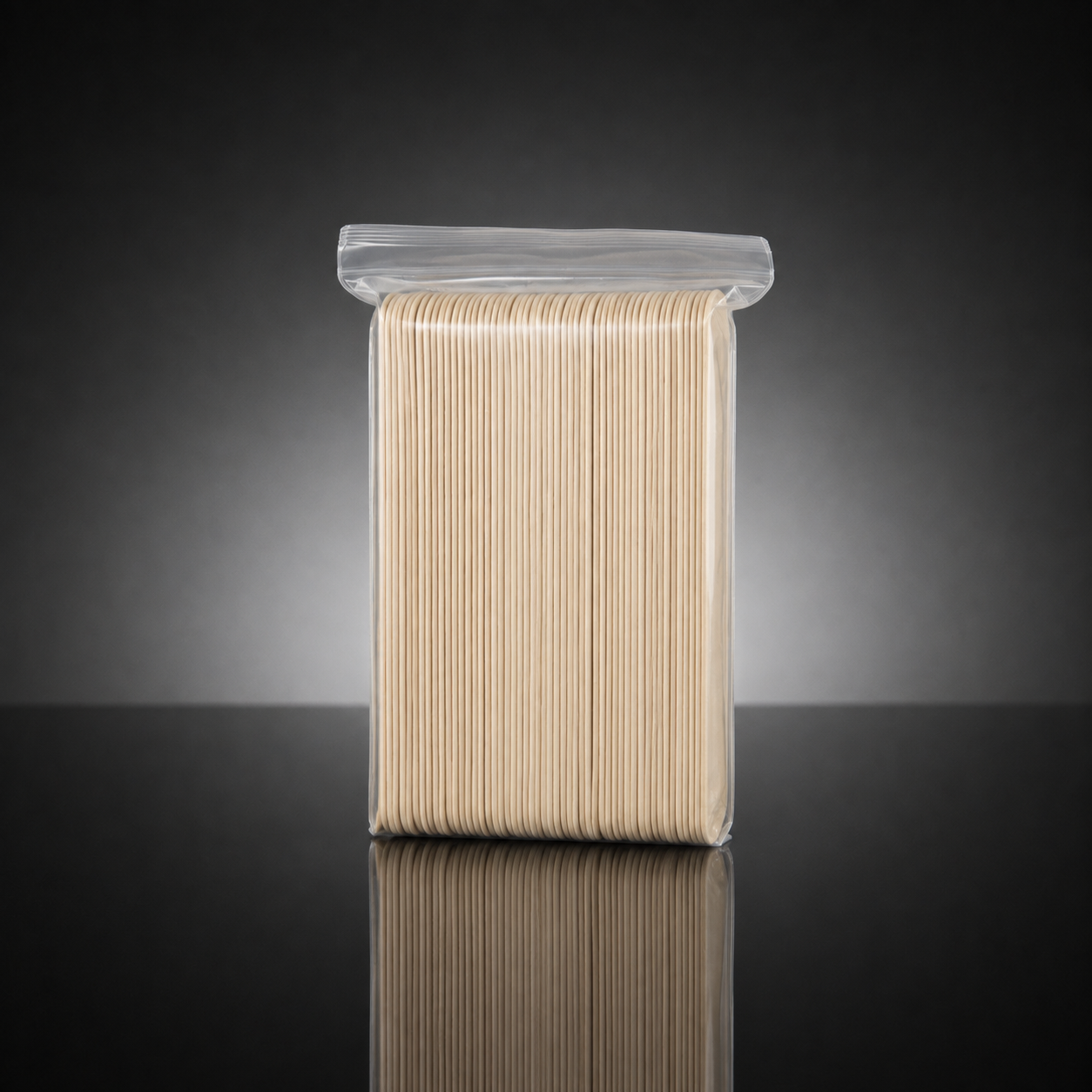 A clear plastic resealable bag containing a bundle of light beige wooden toothpick handles or toothpick-related sticks, photographed upright on a dark reflective surface against a black-to-gray gradient background.