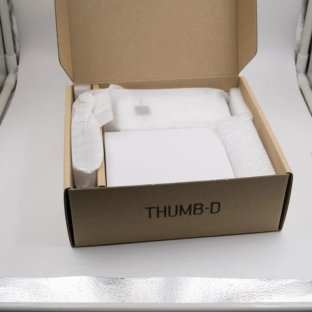 An open brown cardboard box labeled 'THUMB-D' containing white protective packaging materials and a white rectangular item, photographed on a light surface.