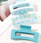 Three hair claw clips arranged diagonally on a white surface. The top clip is light pastel blue, the middle clip is clear with a blue and yellow floral print, and the bottom clip is a solid turquoise blue.
